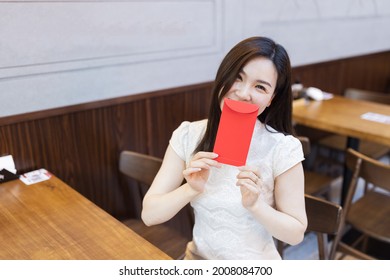Beautiful young asian woman in traditional white dress named cheongsam sitting in Japanese restaurant or cafeteria and waiting ordered food. Girl holding red envelope. Restaurant interior decorated