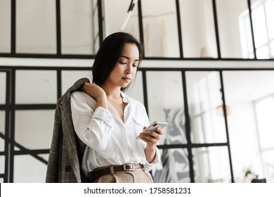 Schöne junge Asiatin, die ein weißes Hemd trägt und in einem modernen Wohnzimmer steht, mit Mobiltelefon