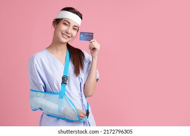 Beautiful Young Asian Woman Wearing Patient Gown And Soft Splint Due To Sore Arm Show Credit Card On Pink Background, Personal Accident Concept.