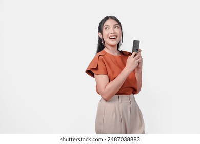 Beautiful young Asian woman wearing brown shirt is holding her smartphone while looking at the copy space above her over isolated white background. - Powered by Shutterstock