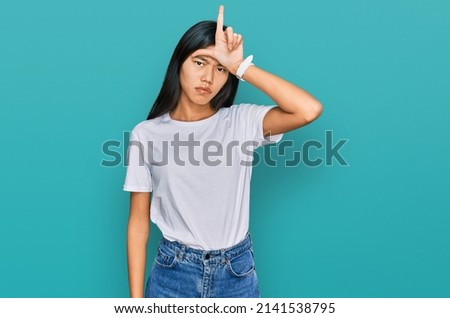 Beautiful young asian woman wearing casual white t shirt making fun of people with fingers on forehead doing loser gesture mocking and insulting. 
