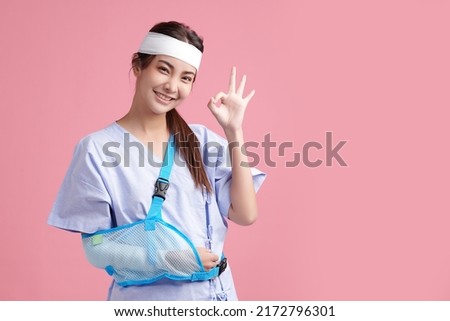 Beautiful young asian woman wearing patient gown and soft splint due to sore arm showing okay sign on pink background, personal accident concept. Stock photo © 