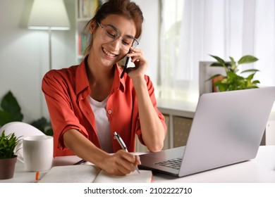 Beautiful Young  Asian Woman Work At Home With Laptop And Talking On Smartphone And Smiling. Female Working On Computer And Using Cell Mobile Phone While Sitting At Home