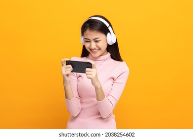 Beautiful young asian women play mobile game and put on wireless headphone standing on isolated yellow background. Playing game on smartphone winning victory moment. Very enjoy and fun relax time