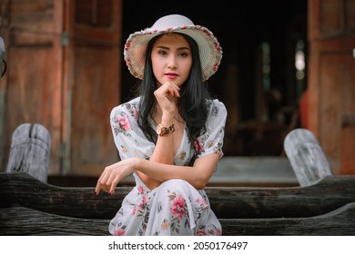 Beautiful young asian women in retro flowers pink dresses styles and hat vintage sitting on wooden stair front view old house background and looking camera, natural light portrait profile in vacation 