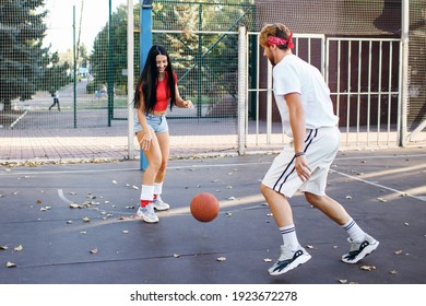 A Beautiful Young Athletic Brunette Is Playing Basketball With Her Boyfriend And Is Trying To Pick Up The Ball. Young People Have Fun And Fitness. Girl In Denim Shorts And A Red T-shirt