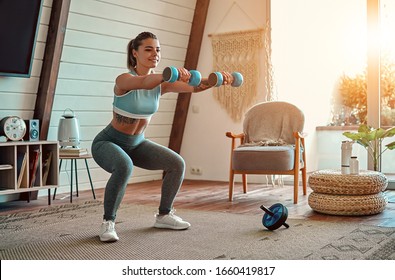Beautiful young athletic girl in leggings and top crouches with dumbbells at home. Sport, healthy lifestyle. - Shutterstock ID 1660419817