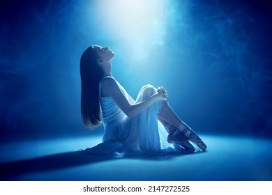 A beautiful young ballerina in a white dress and pointe shoes sits on the stage, hugging her knees, and looks up at the light with hope. Ballet. - Shutterstock ID 2147272525