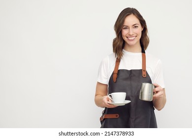Beautiful young barista woman looking in camera and smiling, while serving a cup of coffee and holding a milk jug. Isolated on white background.