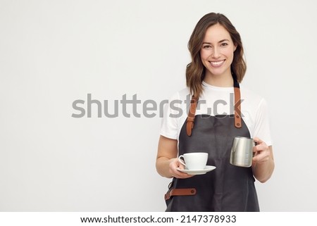 Beautiful young barista woman looking in camera and smiling, while serving a cup of coffee and holding a milk jug. Isolated on white background.
