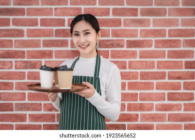 Beautiful young barista woman wearing apron holding tray coffee with ready to serve customer in cafe. Portrait of asian female barista smile and show tray with coffee glass on brick wall background. - Powered by Shutterstock