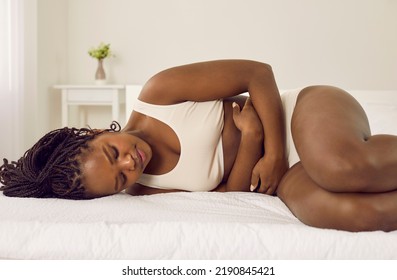 Beautiful Young Black Girl Suffering From Severe Period Cramps. Sad, Stressed African American Woman In Underwear Having Acute Menstrual Pain, Lying On Bed And Holding Hands On Her Belly