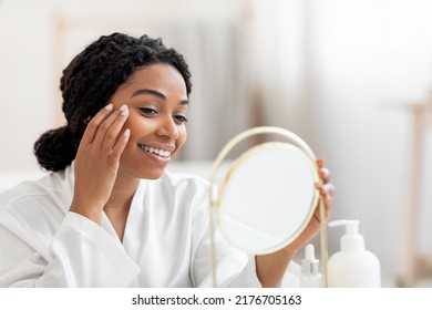 Beautiful Young Black Lady Looking In Mirror And Applying Under Eye Cream, Happy Smiling African American Woman In White Silk Robe Making Beauty Treatments At Home, Enjoying Self-Care, Closeup