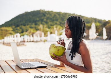 Beautiful Young Black Woman Drinking Refreshing Coconut Water When Sitting On Beach And Reading Article On Laptop Screen