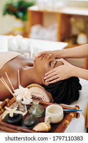 Beautiful Young Black Woman Enjoying Face And Head Massage In Spa Salon When Lying On Bed Next To Tray With Aroma Oils