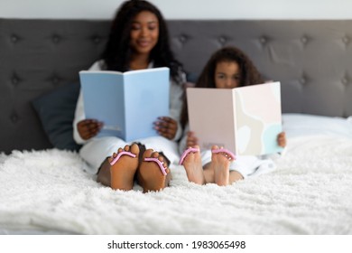 Beautiful young black woman and her cute daughter reading fashion magazines and doing pedicure after bath on bed, selective focus. Domestic beauty spa day for parent and child