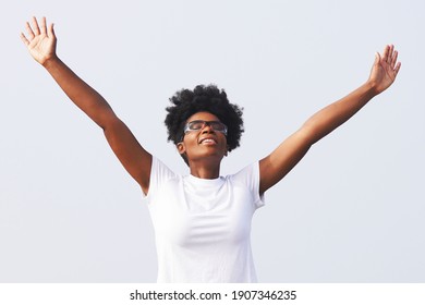 Beautiful Young Black Woman Holding Her Arms Up In The Air To Pray And Celebrate While Smiling And Wearing A White Shirt, Afro Pony Tail And Glasses                              