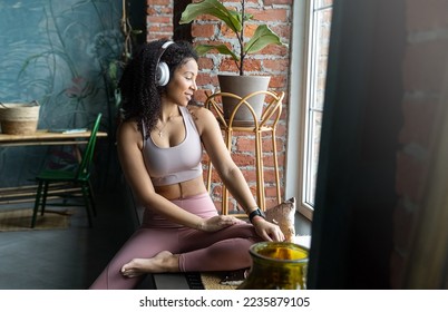 Beautiful young black woman listen music stylish white headphones wear sportswear sitting windowsill dark loft apartment background. Copy space, mock-up,, blank. Concept of music in daily youth life