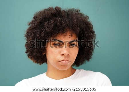 Beautiful young black woman looks confused or angry wearing a white tee shirt and natural curly afro, standing against a teal green background                               