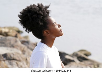 Beautiful young black woman outside sitting at the beach with her eyes closed on the rocks while wearing a white shirt and looking out towards the ocean with the sun on her face                       