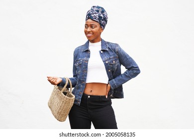 Beautiful young black woman is outside going shopping wearing a jeans jacket and a patterned turban, carrying a basket purse and smiling, looking left                              