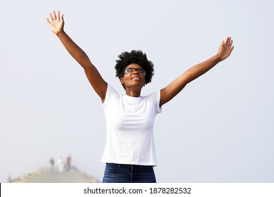 Beautiful Young Black Woman With Protective Eyewear On Looks Up At The Sky With Arms Raised Wearing A White Shirt And Jeans With Palms Outstretched                 