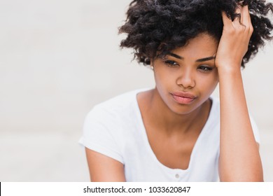 Beautiful young black woman with sad, pensive, reflective look, against white wall background with copy space for your text or advertising content. Portait of worry dark skinned female in white tshirt