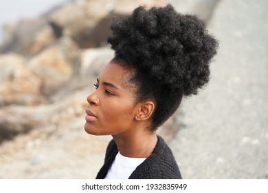 Beautiful young black woman sits at the beach on the rocky shore on a foggy cool morning with her hair up in an afro pony tail looking out at the water                            