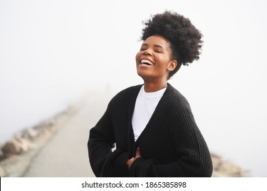 Beautiful young black woman stands on the jetty at the ocean wearing a white shirt and black cardigan sweater with an updo high puff hairstyle, laughing on a foggy day                              