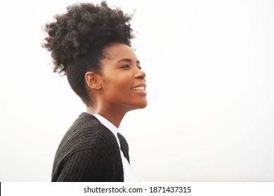 Beautiful young black woman stands against the fog at the beach with a white shirt, black sweater, and updo hairstyle looking to the right, smiling for a portrait                               