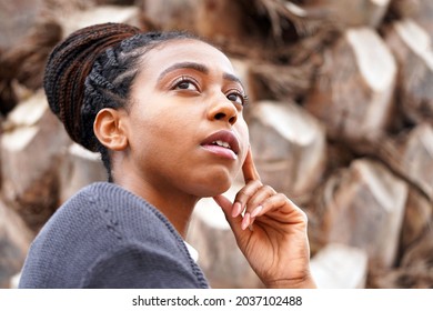 Beautiful Young Black Woman Stands In Front Of Tree Bark Outside And Puts Her Hand To Her Face While Looking Up And Thinking                              