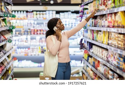 Beautiful Young Black Woman With Tote Bag Making Phone Call While Shopping For Food At Grocery Department Of Big Mall. Lovely African American Lady Purchasing Products At Supermarket