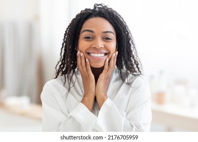 Beautiful Young Black Woman Touching Her Face And Smiling At Camera While Making Beauty Treatments At Home, Attractive African American Female Wearing White Silk Robe Enjoying Selfcare, Copy Space