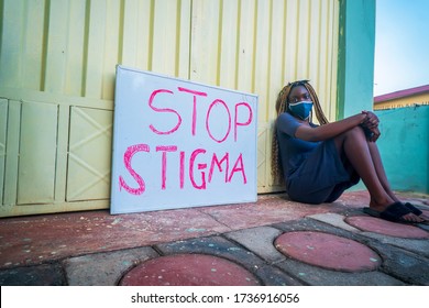 A Beautiful Young Black Woman, Wearing A Locally Made Mask Sitting Beside A Placard Educating On Stop Stigma On People With Corona Virus Disease - Black Millennial Covid-19 And Stigmatisation.