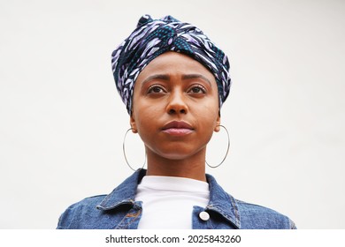 Beautiful young black woman wearing a printed head wrap turban, silver hoop earrings and a jeans jacket looks forward and up against a white background                               