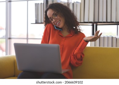 Beautiful Young Black Woman Wearing Orange Shirt Using Laptop And Smartphone For Communication On Sofa At Home. Business Woman And Freelance Work From Home In Spreading Of Coronavirus Covid-19.