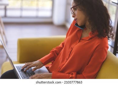 Beautiful Young Black Woman Wearing Orange Shirt Using Laptop And Smartphone For Communication On Sofa At Home. Business Woman And Freelance Work From Home In Spreading Of Coronavirus Covid-19.