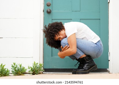 Beautiful Young Black Woman Wearing White Shirt, Jeans And Black Boots, Crouches In Front Of A Teal Door On A White Clapboard House                         