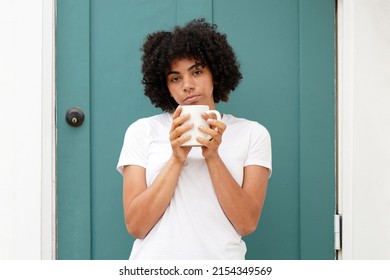 Beautiful Young Black Woman Wearing A White Shirt With A Natural Curly Afro Stands Outside Her Teal Green Front Door Holding A Coffee Mug, Drinking Coffee.                              