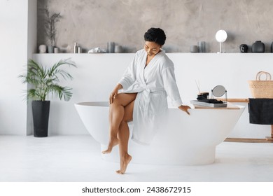 Beautiful young black woman wearing white bathrobe sitting on bathtub and touching her beautiful legs, happy female enjoying silky soft skin after morning shower, posing in modern bathroom interior