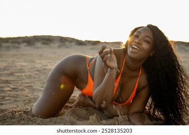 Beautiful young black woman wearing an orange bikini is kneeling on the sand on a beach at sunset playing with the sand - Powered by Shutterstock