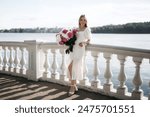 Beautiful young blond girl in white summer dress holding a bouquet of white and pink peonies near a lake. Pretty teenage girl with long blond hair walking on promenade in Europe town. Traveling Europe