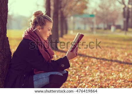 Beautiful young blond leaning on a tree on an autumn day, reading a book