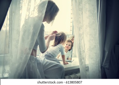 Beautiful young blond mother is braiding hair to her cute little daughter sitting on the window sill in front of a mirror. Image with selective focus and toning - Powered by Shutterstock