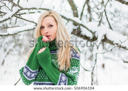 Beautiful young blond woman is dressed in a warm green sweater with Norwegian patterns on the snowy background in winter forest. Girl blowing on hands to warm up. Christmas theme