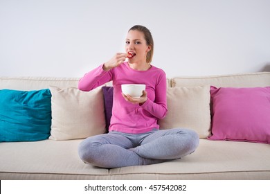 Beautiful Young Blond Woman Sitting On A Sofa At Home, Smiling And Eating Healthy Strawberries Fruit Food In Living Room, House Interior. 