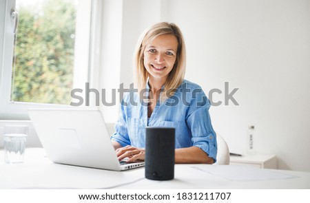 beautiful young and blond woman is talking to a smart speaker and is working at home
