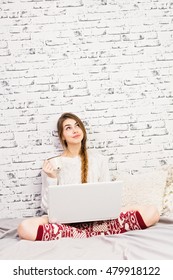 Beautiful Young Blonde Caucasian Woman At Home In Bed, Using Laptop, Holding Coffee Cup, Looking Up, Sitting Cross-legged. Winter Outfit. Brick Wall Background. Copy Space.