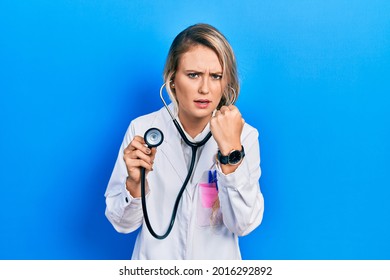 Beautiful Young Blonde Doctor Woman Holding Stethoscope Angry And Mad Raising Fist Frustrated And Furious While Shouting With Anger. Rage And Aggressive Concept. 