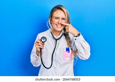 Beautiful Young Blonde Doctor Woman Holding Stethoscope Pointing With Hand Finger To Face And Nose, Smiling Cheerful. Beauty Concept 